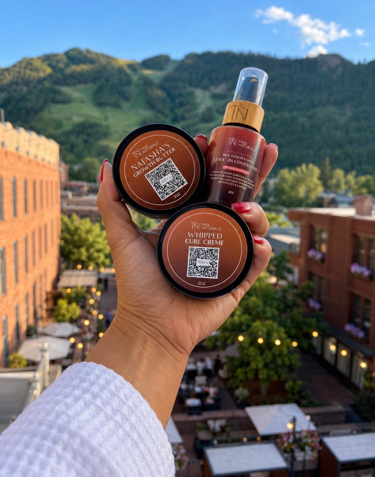 A hand holding the Mini Tree-O Kit: 2 oz Leave-In Conditioner, Whipped Curl Creme and Natasha's Growth Butter, with buildings and mountains behind.