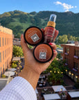 A hand holding the Mini Tree-O Kit: 2 oz Leave-In Conditioner, Whipped Curl Creme and Natasha's Growth Butter, with buildings and mountains behind.