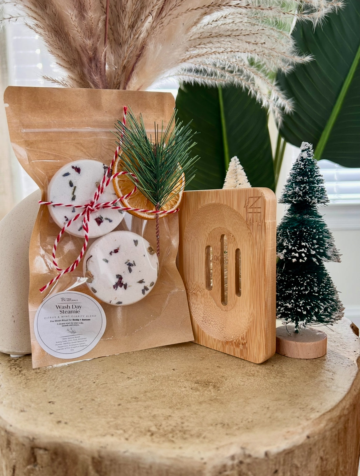 Tree Naturals Wash Day Steamies deluxe duo with bamboo tray styled on a wood pedestal with holiday greenery and mini Christmas tree.