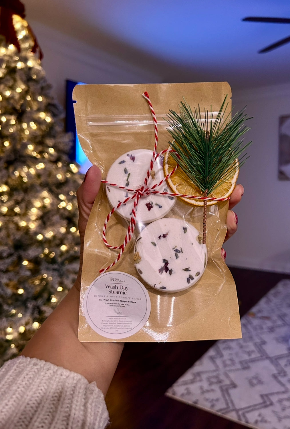 Hand holding Tree Naturals Wash Day Steamies holiday pouch in front of a glowing Christmas tree, showing them as an easy gift.